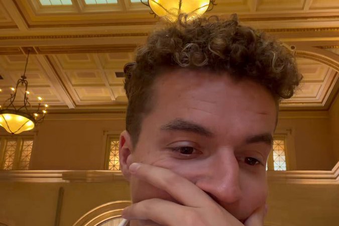 A wedding guest films his own stunned reaction, hand over his mouth, beneath the chandeliers of an ornate reception hall — the opening frame of the AI wedding toast video.