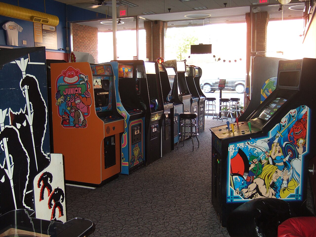 A row of upright arcade cabinets along a carpeted wall in a daylit arcade, screens lit, a bar stool pulled up to one of them
