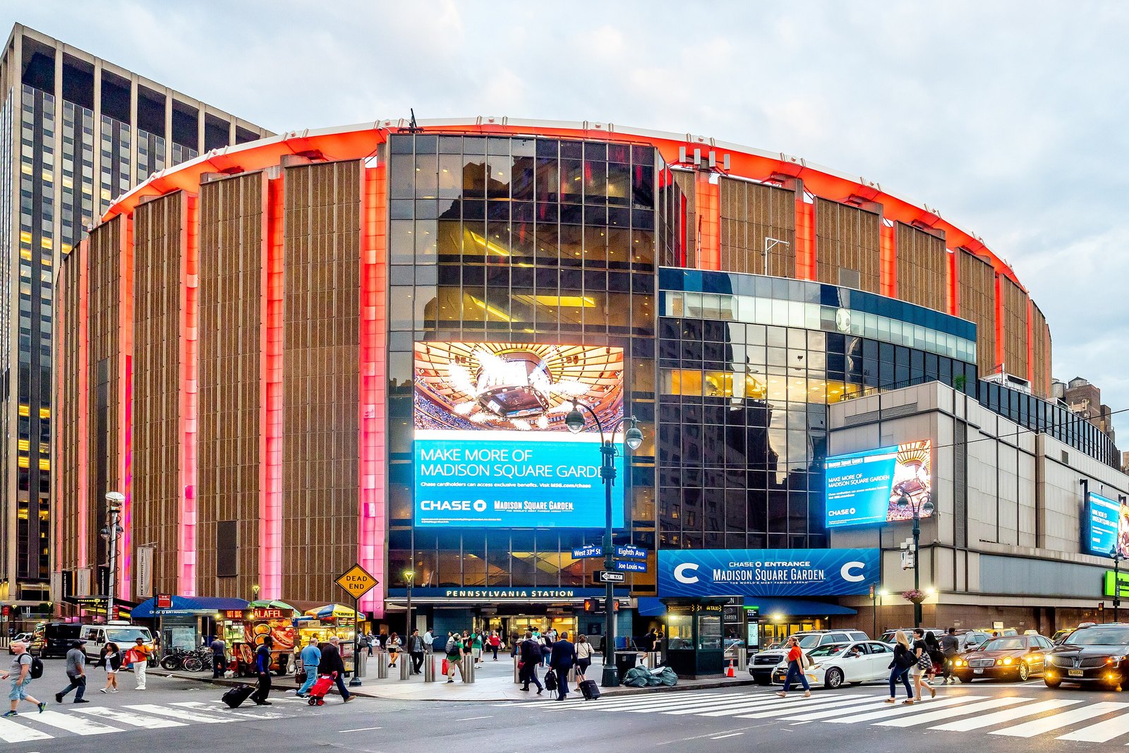 Madison Square Garden at dusk, its curved exterior lit red, with a large blue Chase advertisement on the glass facade and people crossing the street in front.