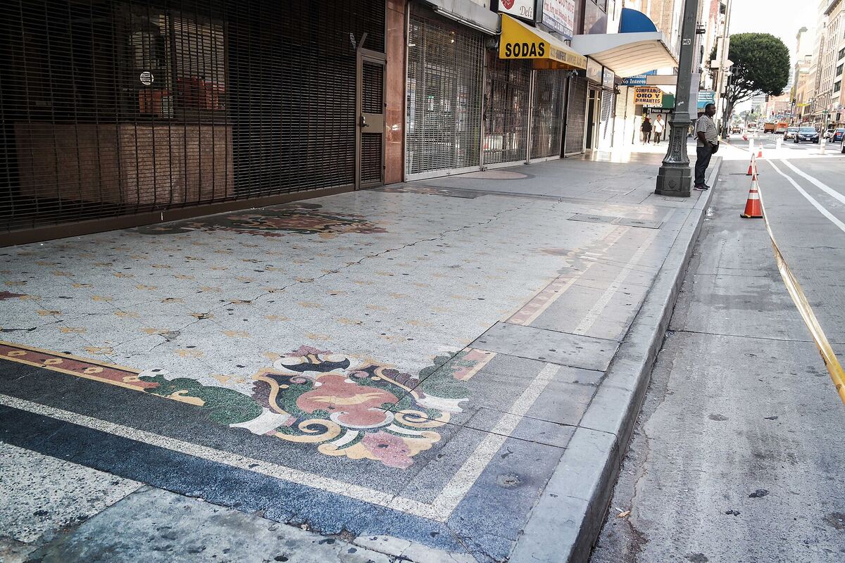 The inlaid terrazzo forecourt of a 1931 movie palace on a downtown Los Angeles sidewalk, its ornamental floral medallions running up to shuttered steel security grilles, with a pedestrian and traffic on the street beyond