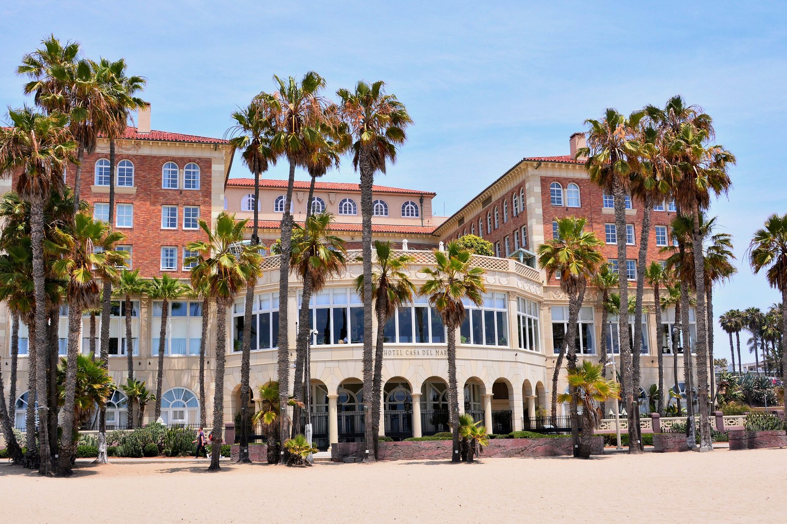 Large multi-storey brick and cream hotel with arched colonnade at ground level, tall palm trees in front, sandy beach below, blue sky above. The name Hotel Casa del Mar is visible on the facade.