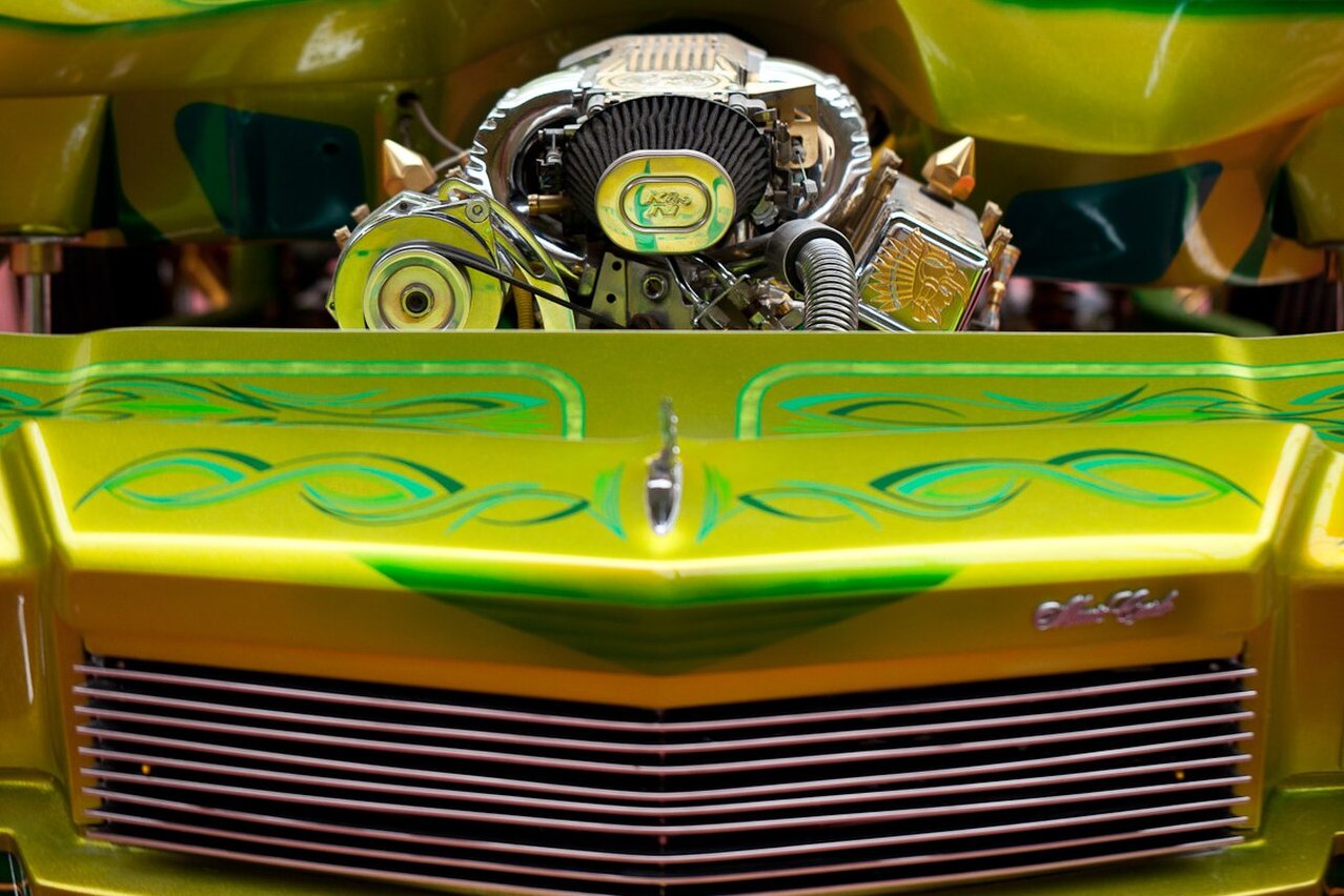 The raised hood of a customized 1979 Chevrolet Monte Carlo, a chromed engine bay above the car's grille, the bodywork in candy green and yellow with airbrushed pinstriping.