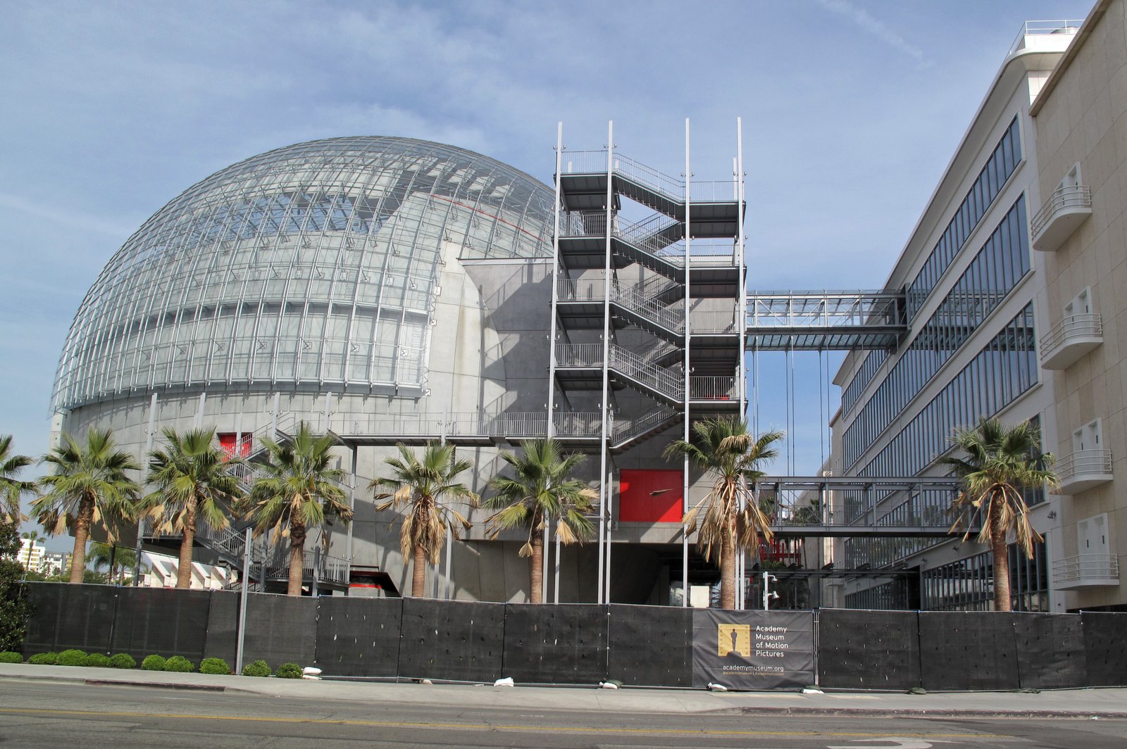 The glass-domed Sphere of the Academy Museum of Motion Pictures, with exterior stair towers, walkways, palm trees, a black construction fence and an Academy Museum banner, seen from the street.