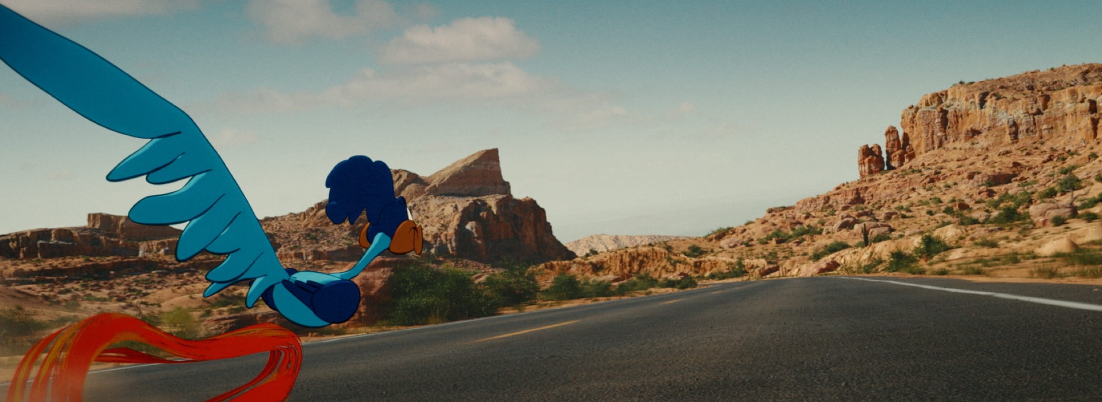 The Road Runner, drawn flat in blue and purple with an orange speed trail, running along a photographed two-lane desert highway past red sandstone mesas in afternoon light — a frame from Coyote vs. Acme.