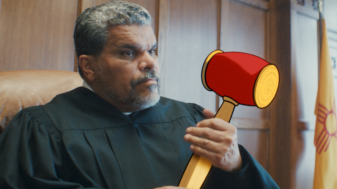 Still from Coyote vs. Acme: Luis Guzman as Judge Troppogrosso in black robes at the bench, straight-faced, raising an oversized red cartoon gavel, with the New Mexico state flag behind him.