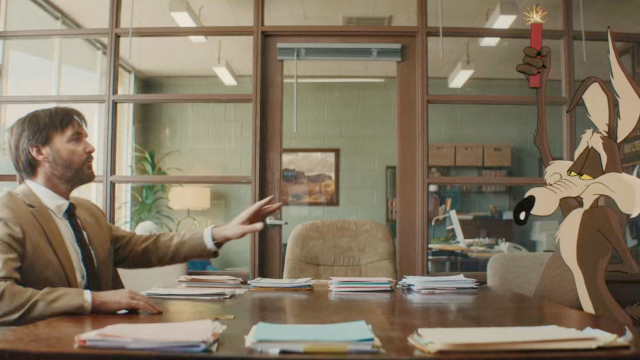 Still from Coyote vs. Acme: Will Forte as lawyer Kevin Avery leans back at a conference table in a strip-mall law office as Wile E. Coyote stands on the table holding a lit stick of Acme dynamite.