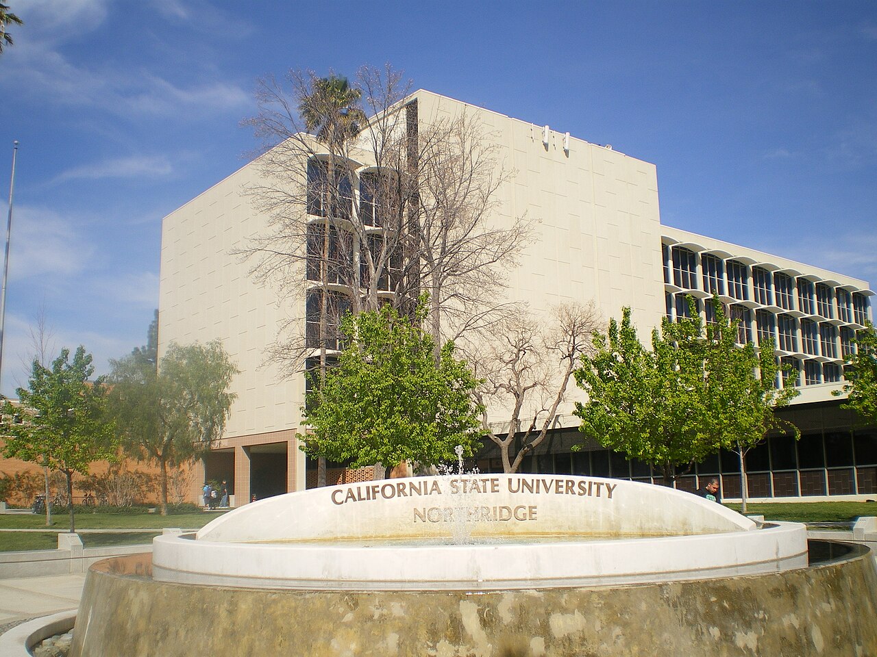 The fountain at Cal State Northridge with the university's name carved into its rim, in front of a white campus building with an arcaded facade under a clear sky
