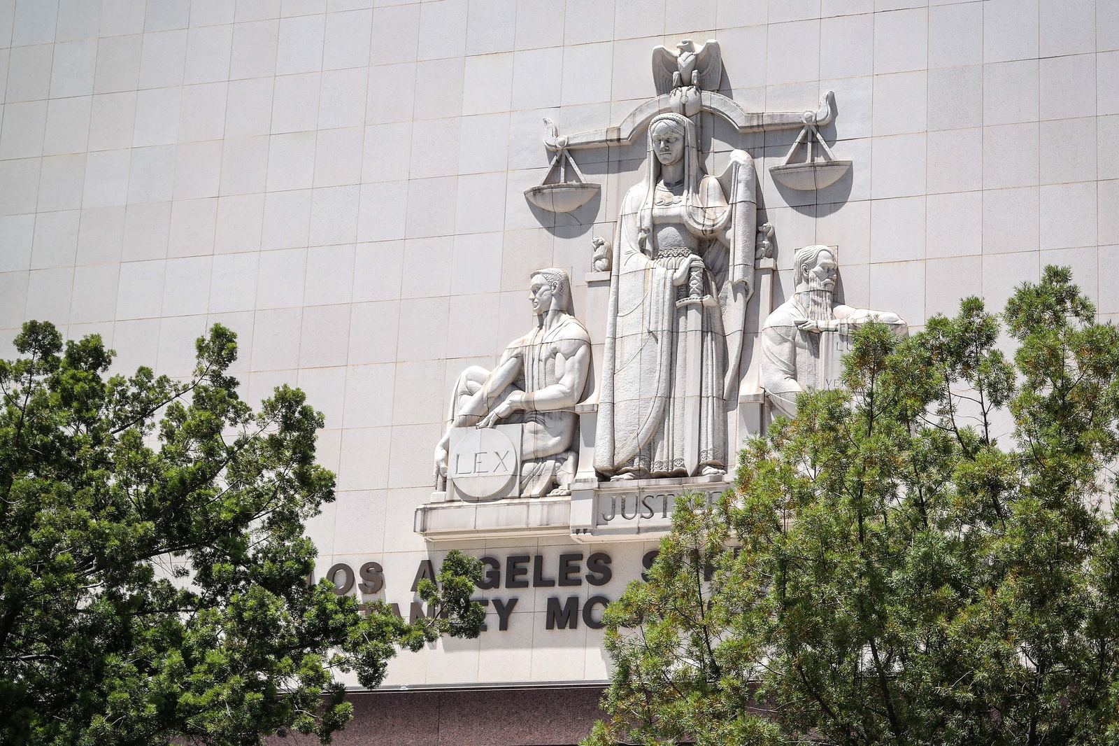 The bas-relief sculpture group on the Hill Street facade of the Stanley Mosk Courthouse in downtown Los Angeles: a robed central figure beneath a set of scales and an eagle, flanked by two seated figures, one resting a hand on a tablet reading LEX, with the building's carved lettering and treetops below