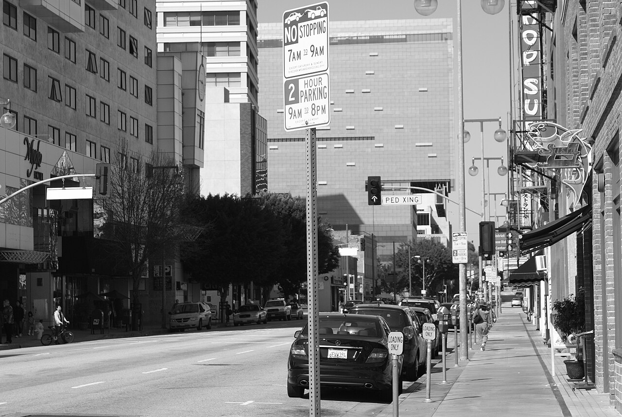 East First Street in Little Tokyo in black and white, a parking sign in the foreground, a chop suey neon blade sign over the sidewalk on the right and low pre-war storefronts running toward downtown towers