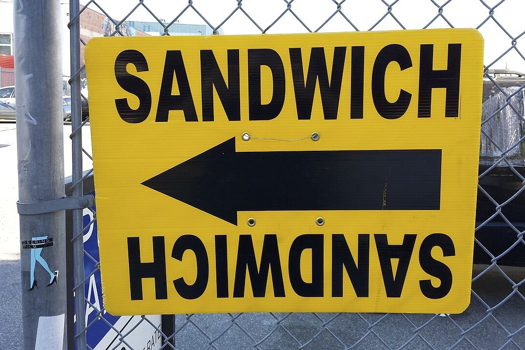 A yellow filming sign reading SANDWICH, with a black arrow pointing left, fixed to a chain-link fence beside a street in the Arts District of Los Angeles.