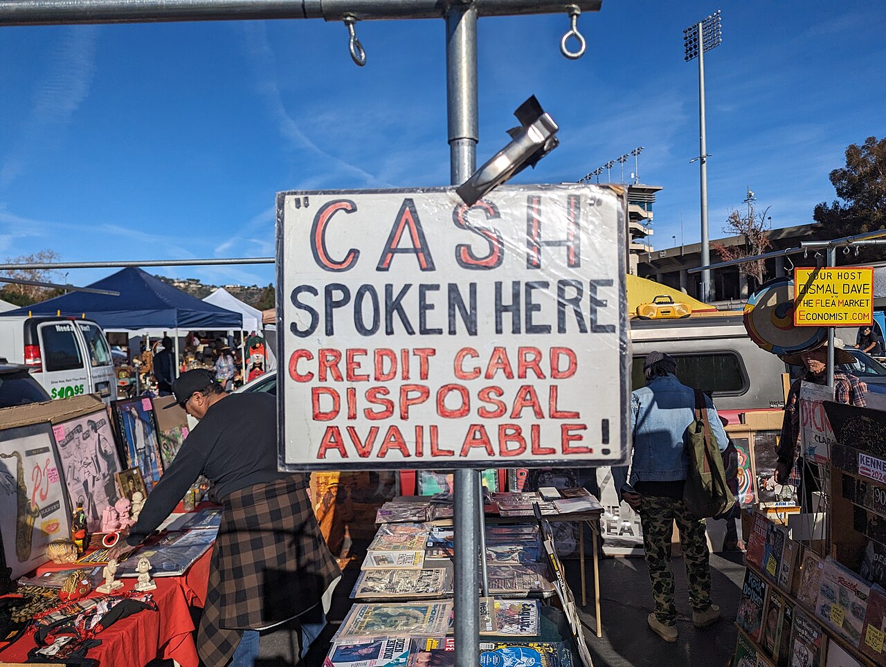 A hand-lettered sign on a pole at a flea market reading "CASH" SPOKEN HERE, CREDIT CARD DISPOSAL AVAILABLE, with tables of old magazines, records and prints behind it and stadium light towers above