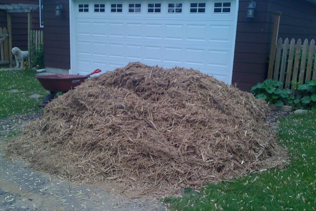A large heap of wood chips tipped onto a driveway in front of a garage, with a wheelbarrow standing at the left and a picket fence and lawn around it