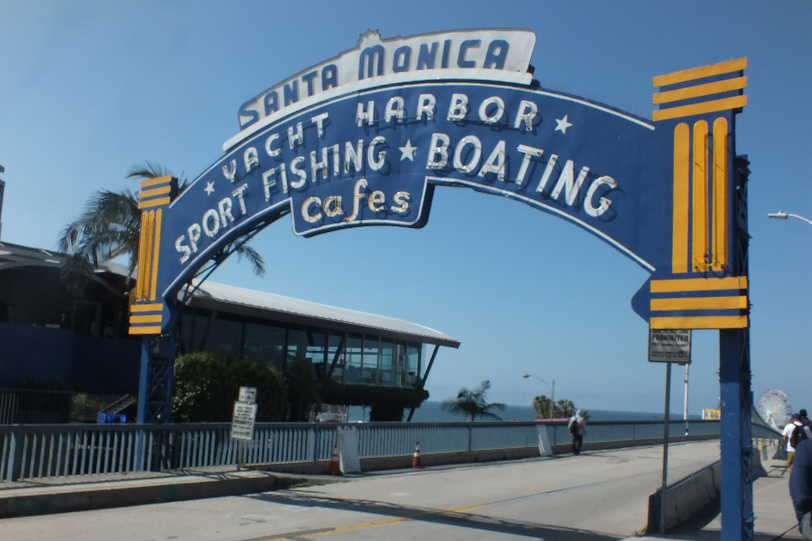 Blue arched entrance sign reading Santa Monica Yacht Harbor Sport Fishing Boating cafes over a pier walkway, with palm trees, a glass building, and the ocean in the background under a clear sky.