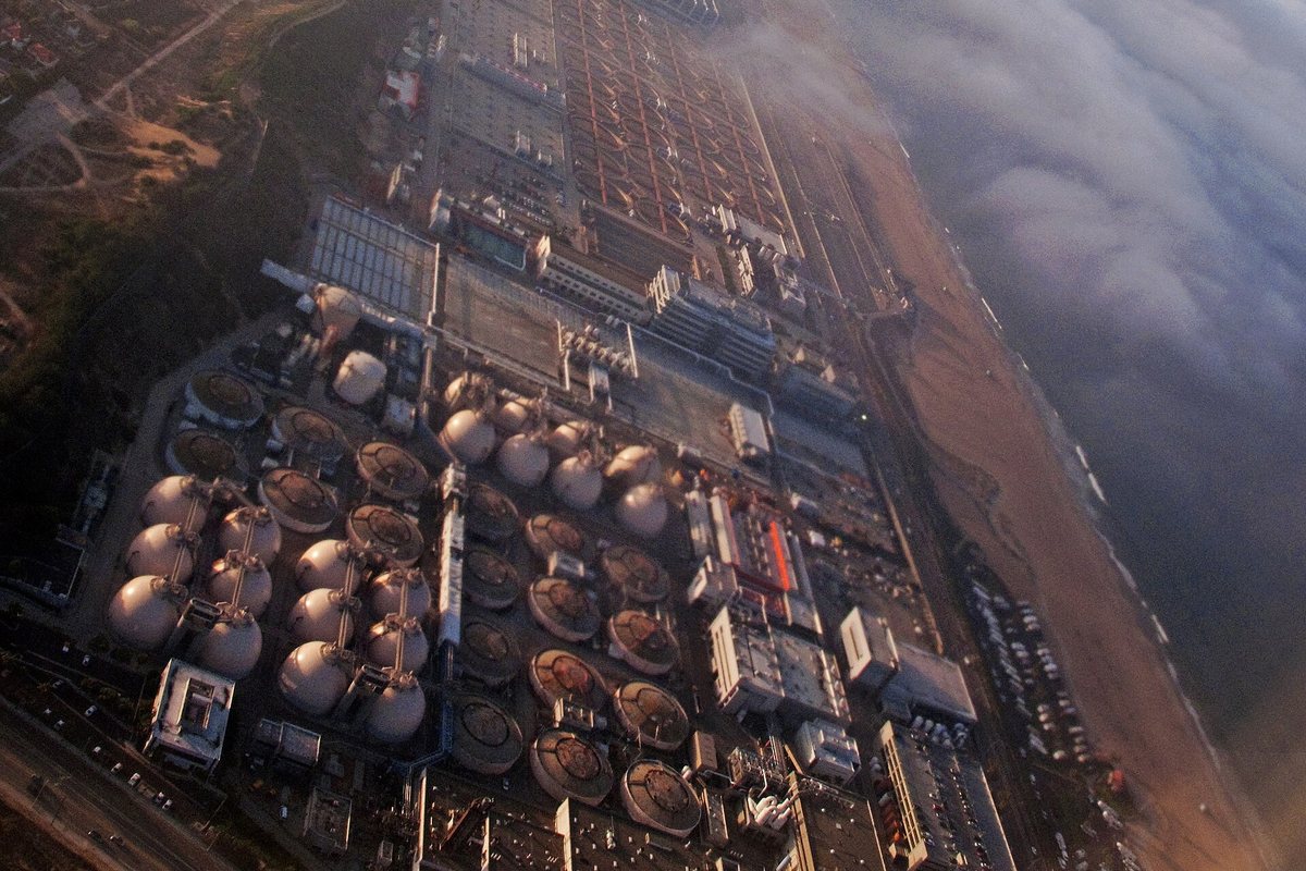 Aerial view of the Hyperion plant's digesters and circular settling basins beside the beach, with the marine layer rolling in offshore.