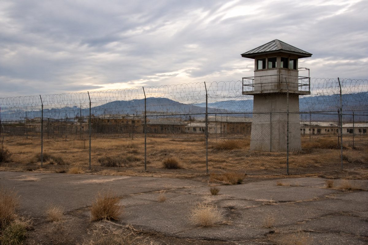 Empty guard tower and chain-link perimeter fence at a closed California prison, dry brush in the foreground and mountains behind