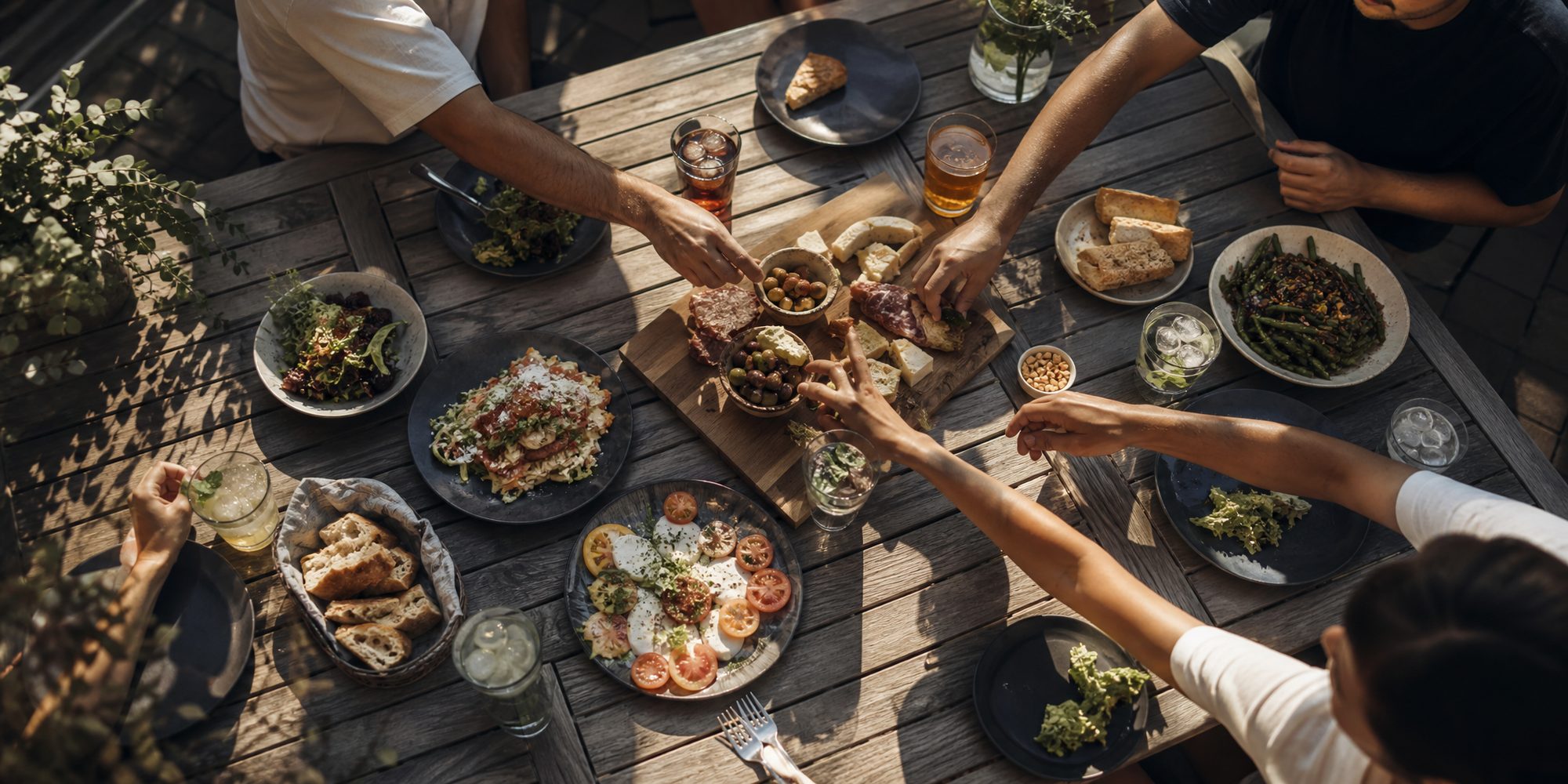 Overhead view of a patio table in late-afternoon light with shared plates, cold drinks and hands reaching across.