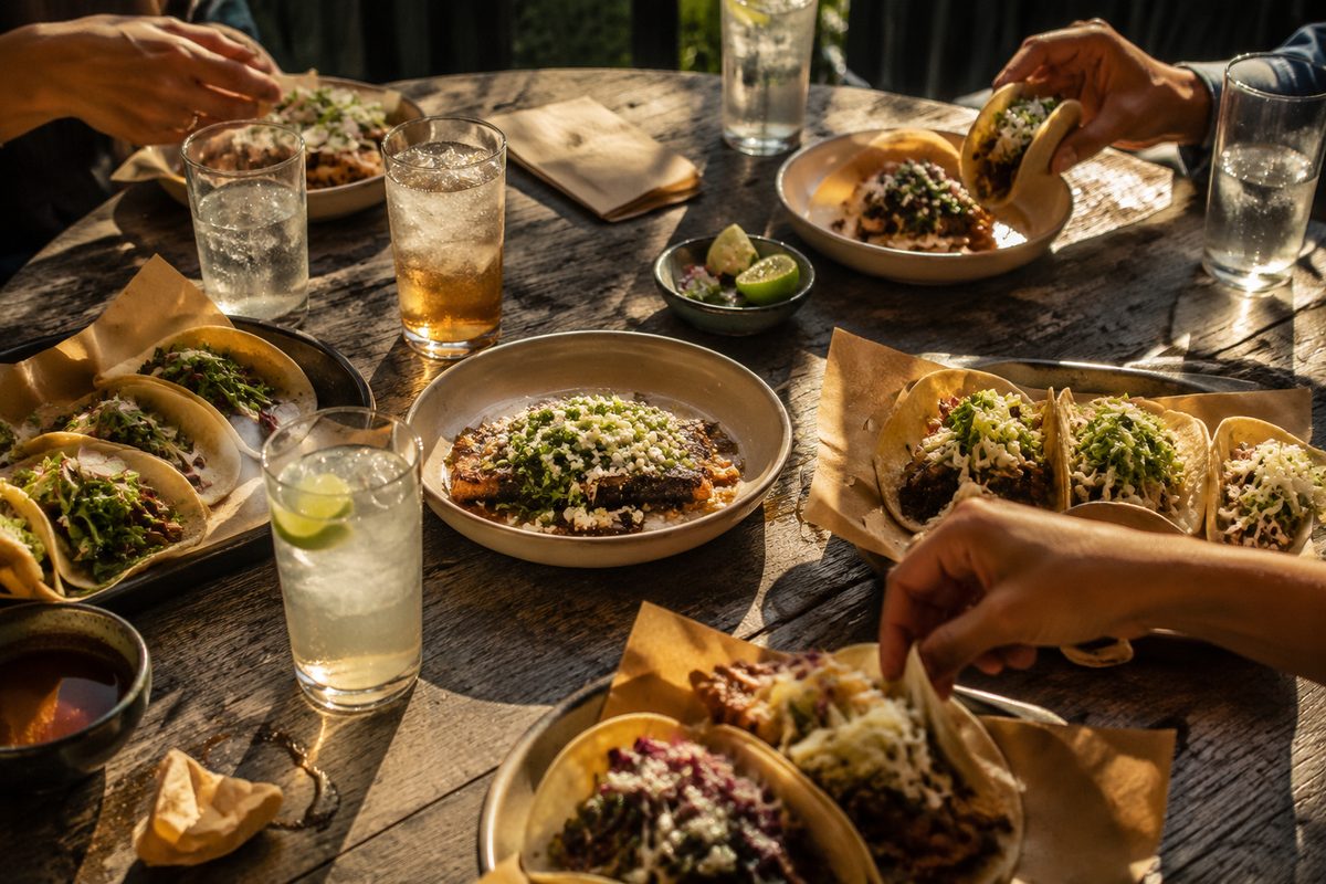 Overhead view of a patio table in late-afternoon light with shared plates, cold drinks and hands reaching across.