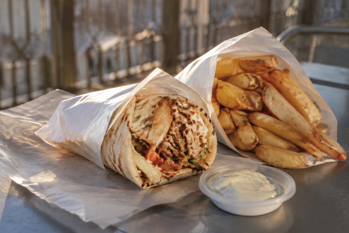 A paper-wrapped shawarma and a paper cone of fries on a metal patio table in late-afternoon light, shot from a three-quarter angle, no branding visible.