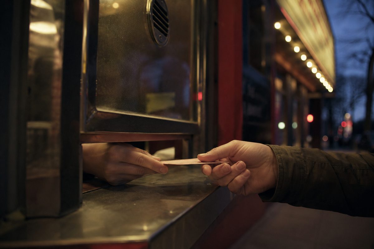 A cinema box-office window at dusk, hands passing a plain paper ticket under the glass, with blurred marquee bulbs and unreadable showtime lettering behind.