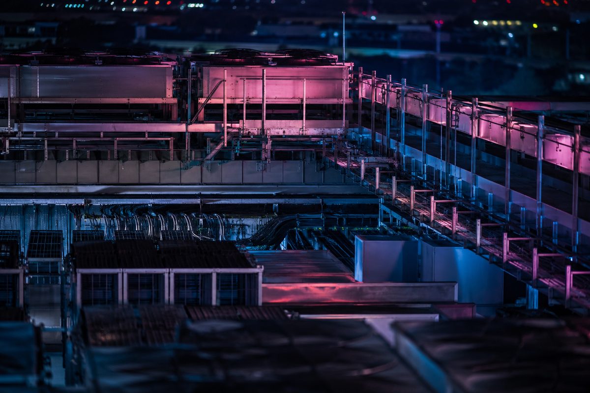 Long-lens night view of stacked rooftop cooling units and cable trays on a data center, lit by distant neon.