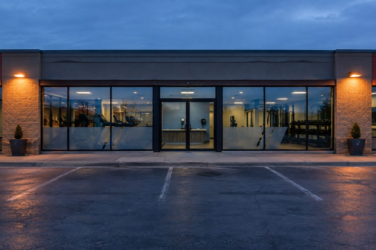 A low-slung gym storefront seen straight on at dawn, glass doors dark, an empty parking lot in front.