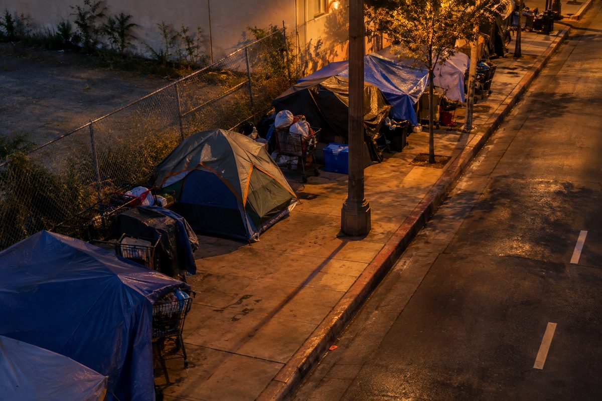 An overhead view at dusk of makeshift tents and tarps lining a wide city sidewalk, lit by orange streetlight.