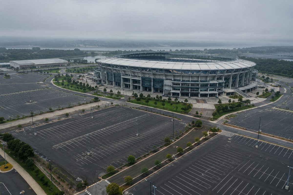 Overhead view of empty striped parking rows, light poles and fresh asphalt near a large stadium under low morning fog.