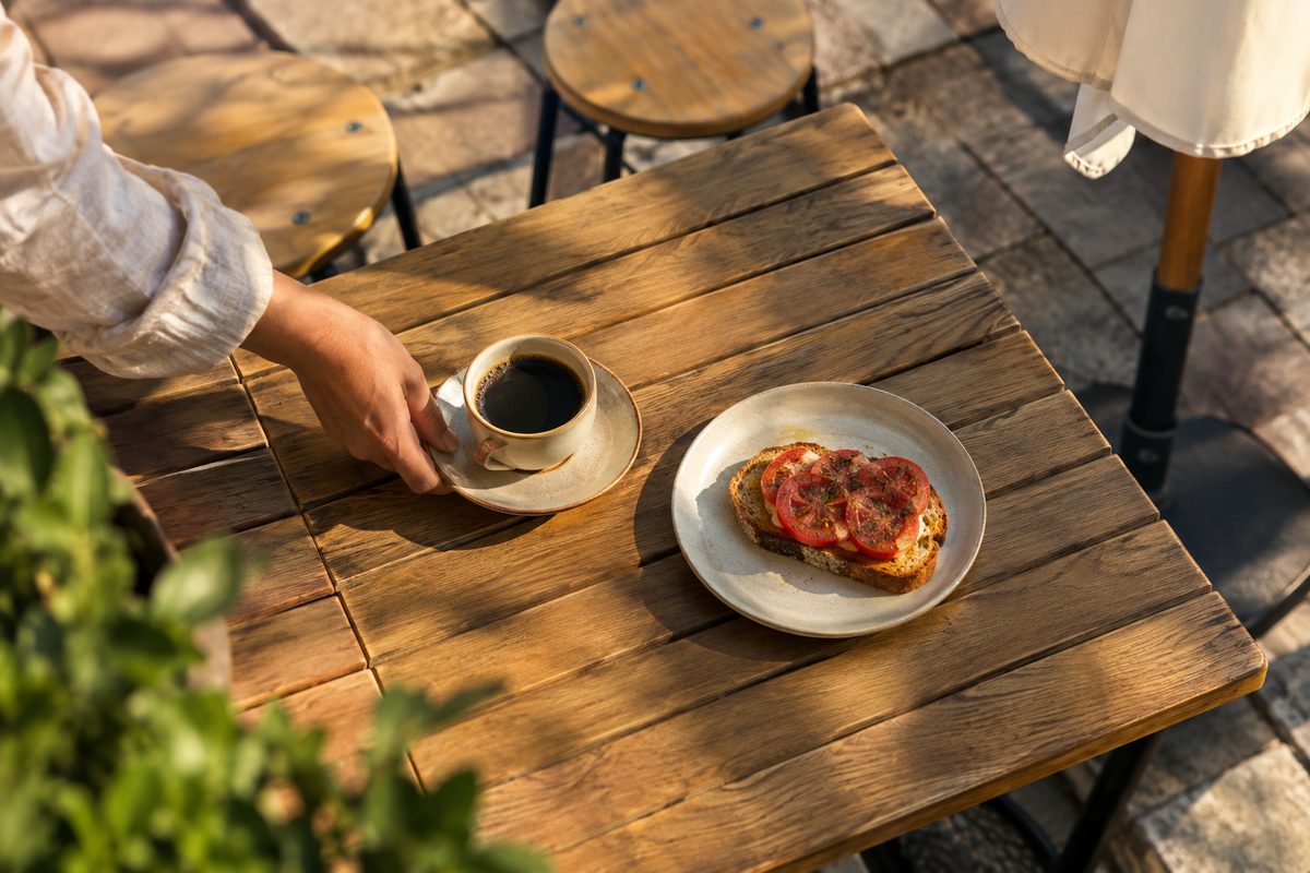 Overhead view of a coffee cup and a slice of toast on a wooden outdoor platform table in late-day light, with empty chairs behind.