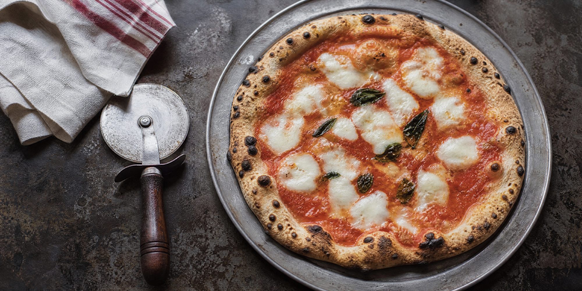 Overhead view of an unbranded Neapolitan-style pizza with a charred, blistered rim on a steel pan, next to a cutting wheel and a folded cloth.