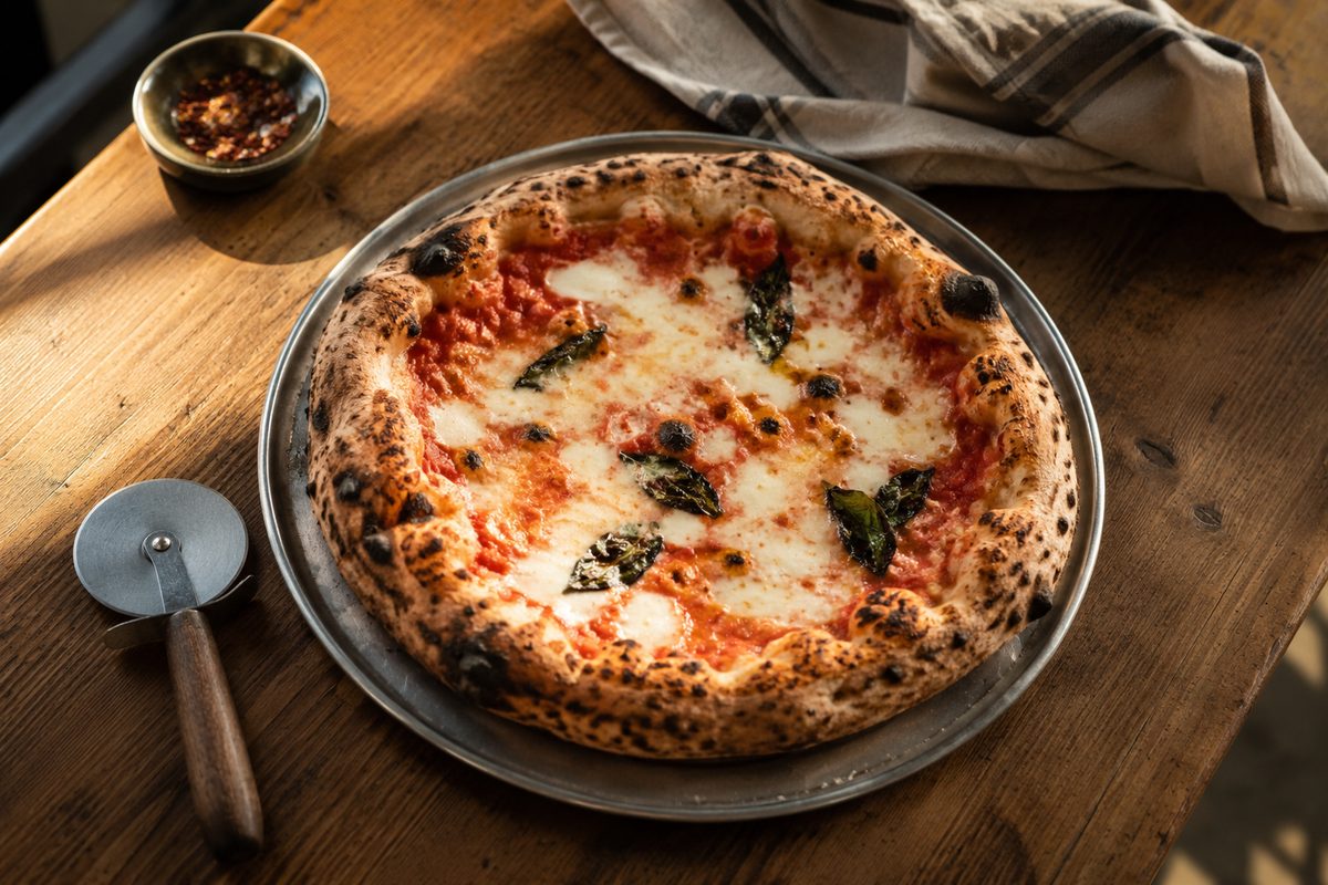 Overhead view of an unbranded Neapolitan-style pizza with a charred, blistered rim on a steel pan, next to a cutting wheel and a folded cloth.