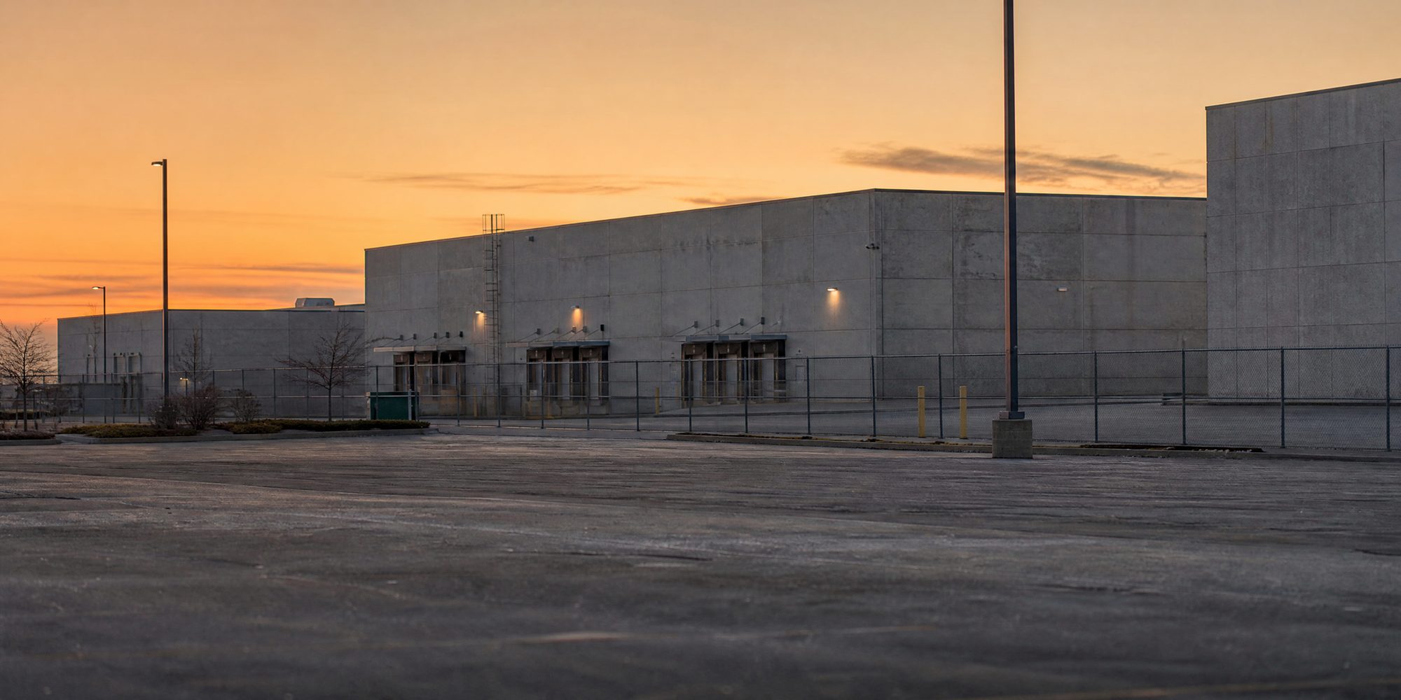 A long-lens view across an empty parking lot at dawn toward a row of windowless concrete industrial buildings with loading docks and chain-link fencing, no signage visible.