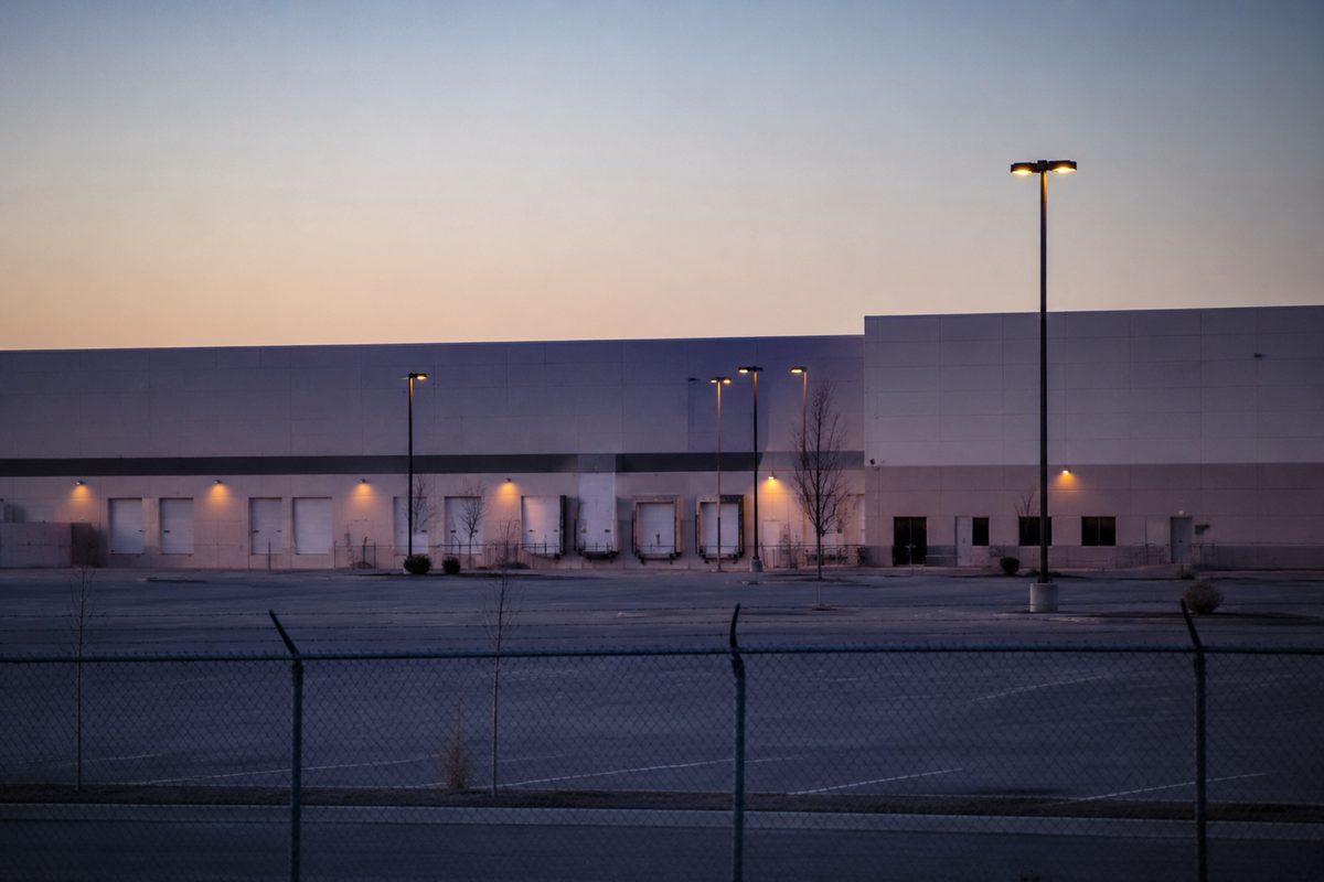 A long-lens view across an empty parking lot at dawn toward a row of windowless concrete industrial buildings with loading docks and chain-link fencing, no signage visible.