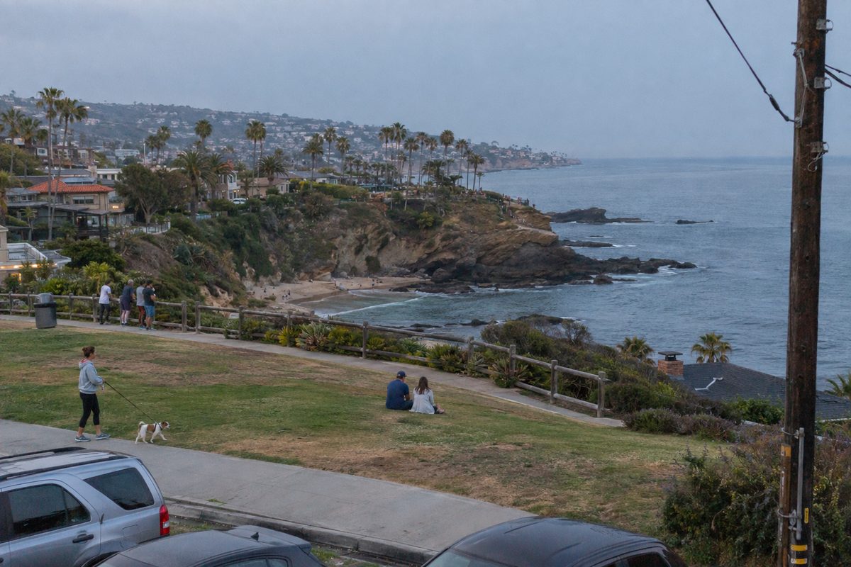 Early-evening view of the grassy clifftop at Crescent Bay Point Park in Laguna Beach, with people at the railing above the ocean and a woman walking a dog along the path.