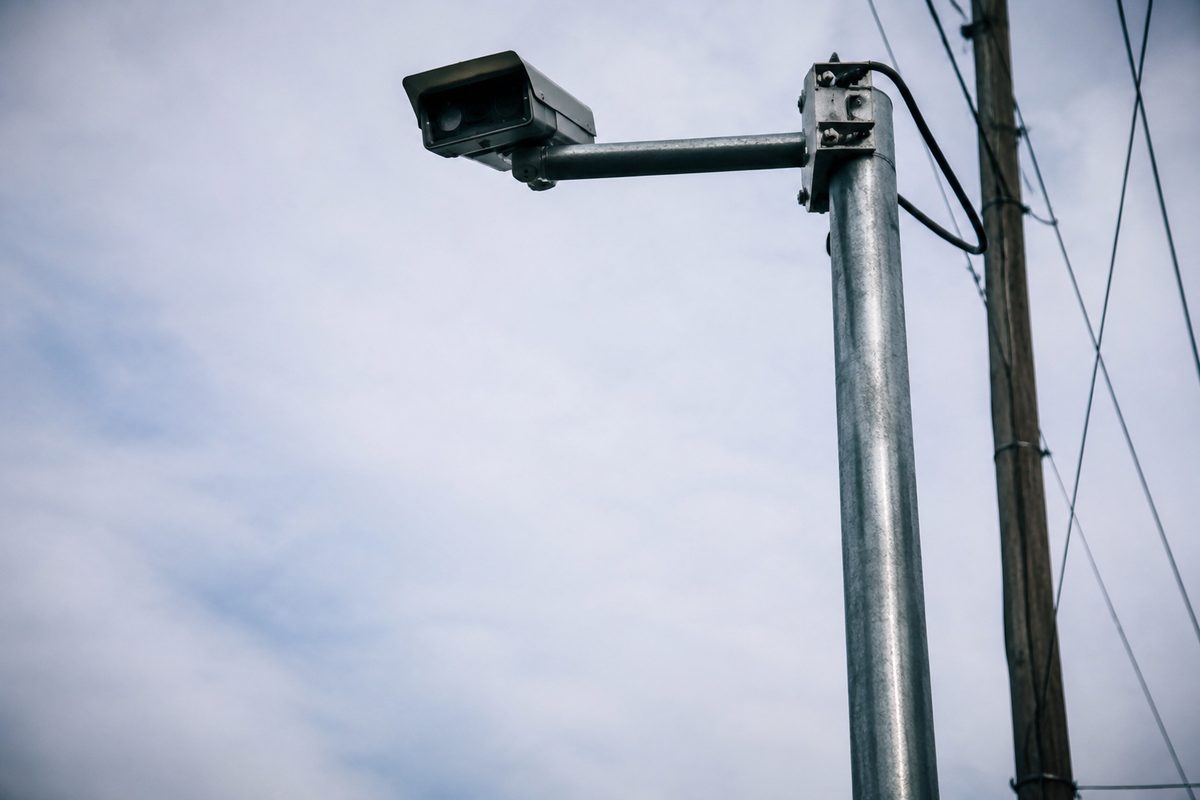 A pole-mounted traffic camera seen from below against a pale open sky, wires running down the pole.
