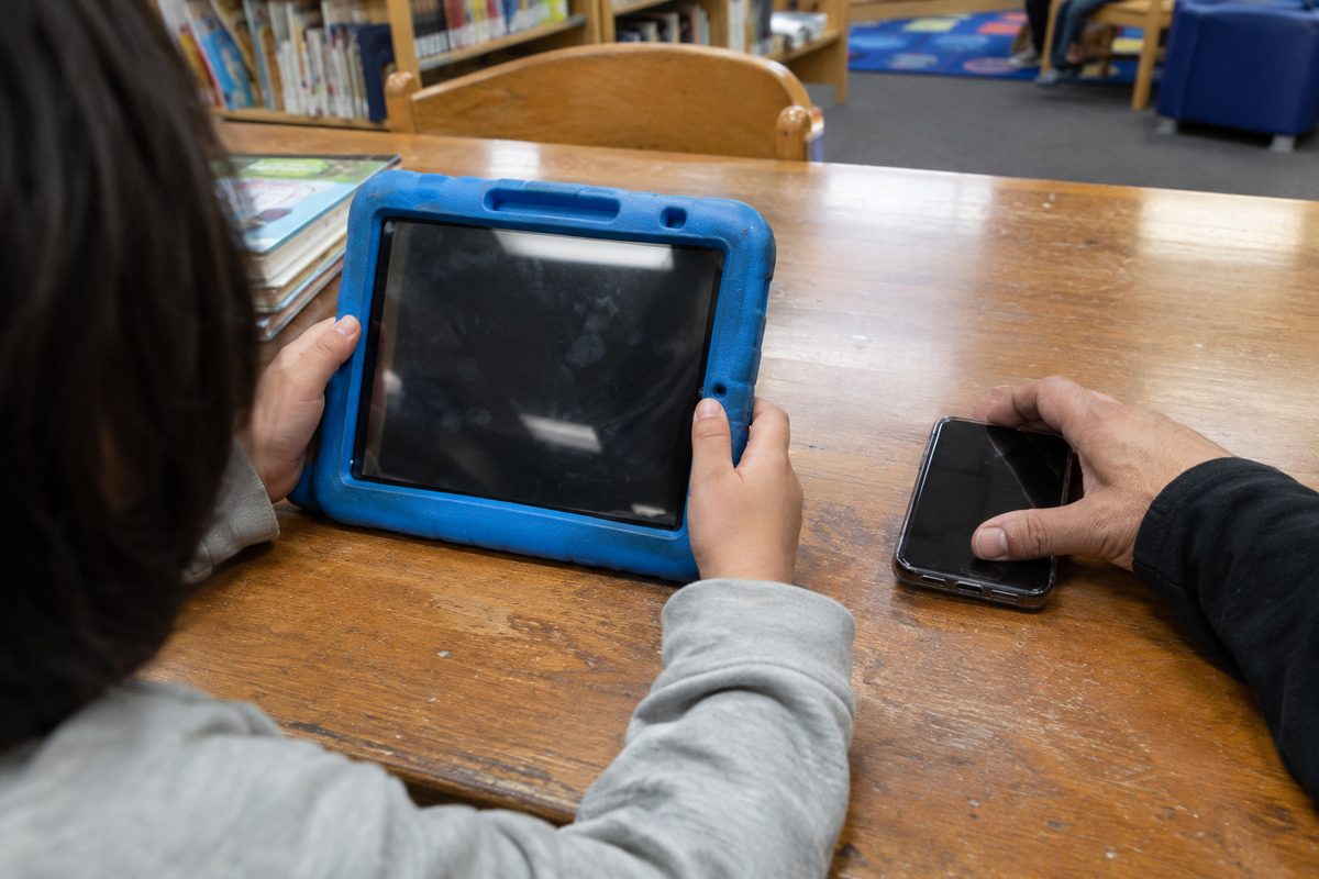 A child's hands hold a tablet in a thick foam bumper case on a library table while a parent's hand rests nearby holding a phone.