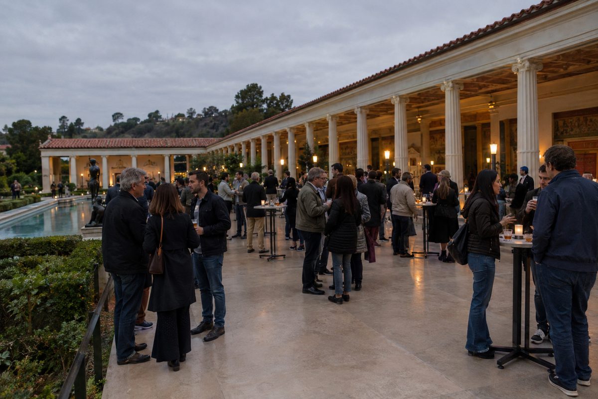 Adults holding small glasses of amber beer at tables on the Getty Villa's café terrace at dusk, with Roman-style columns behind them.