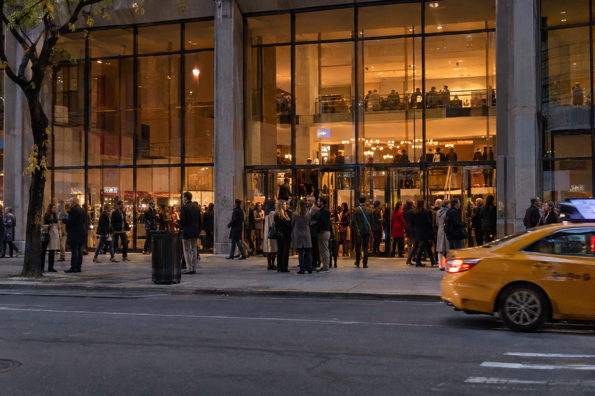 Festivalgoers gather under the lit glass lobby of a Lincoln Center theater on Manhattan's Upper West Side at night, seen from across the street.