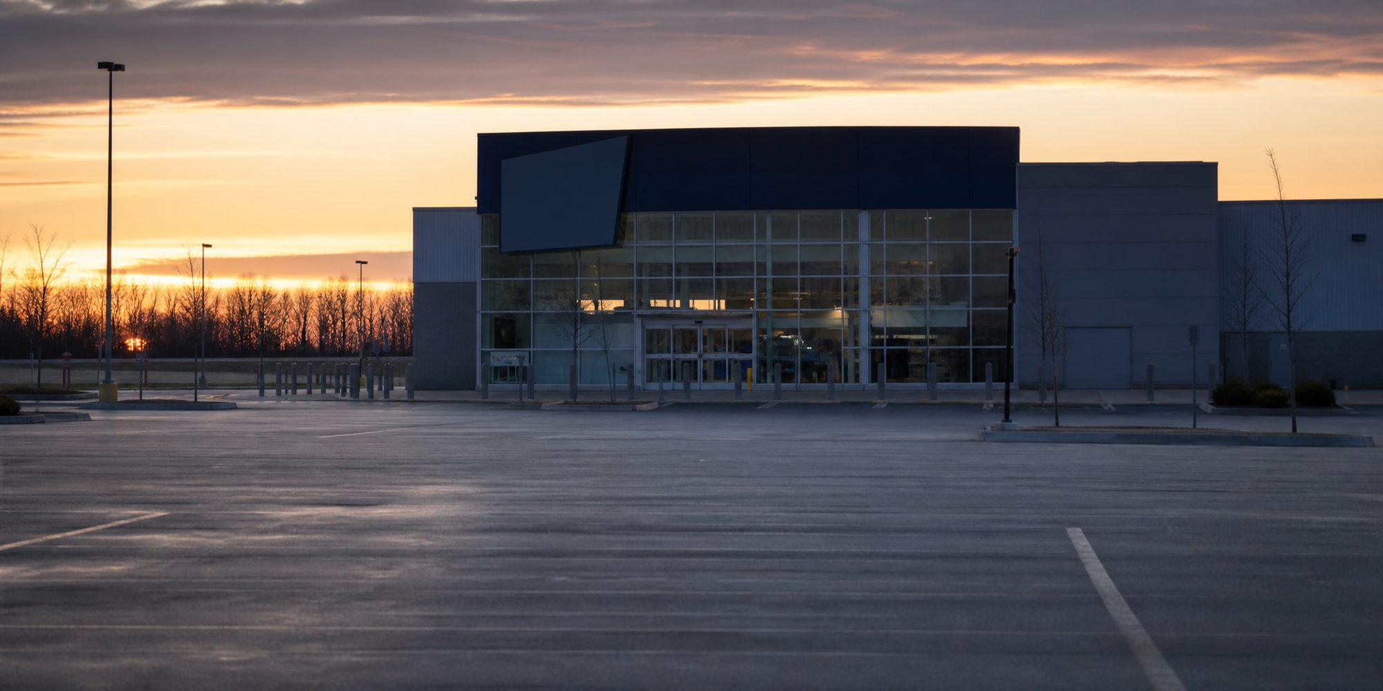 A long-lens view across an empty parking lot at dawn toward an anonymous glass-fronted electronics store with unreadable signage.