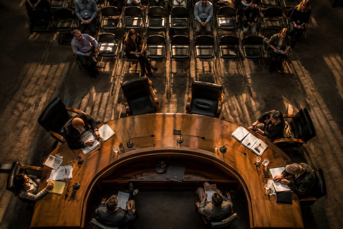 Overhead view of a public hearing room at dusk, a horseshoe table with several empty seats, agenda papers and water cups, a few seated figures seen from above.