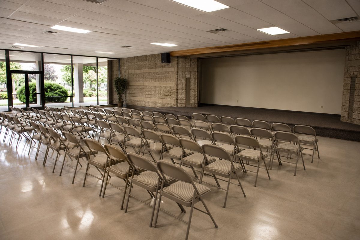 An empty community meeting hall with rows of folding chairs facing a low stage, no people present
