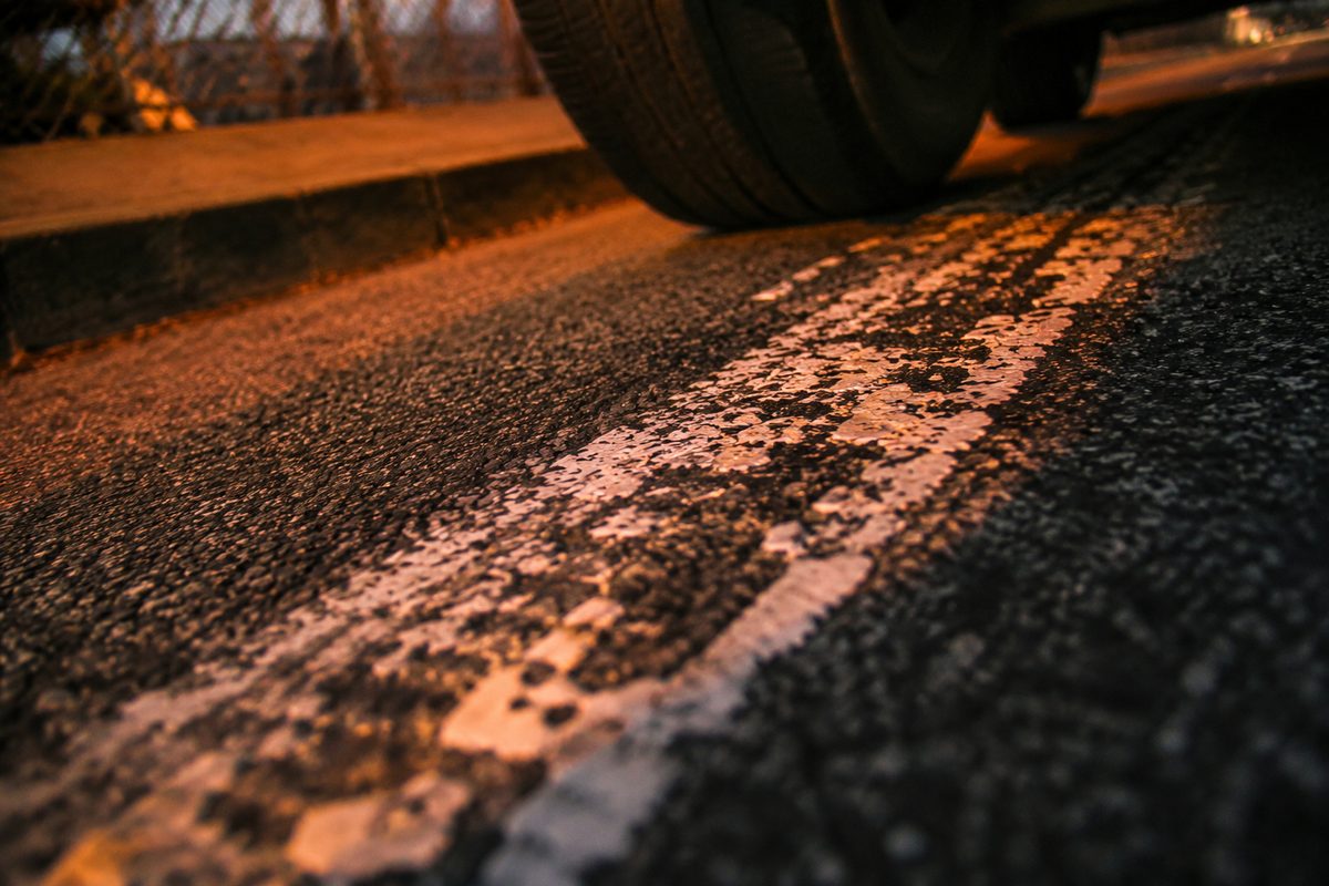 Close-up of a worn white lane stripe on dark asphalt at dusk, reflective paint beads catching orange streetlight, a car tire at the edge of the frame.