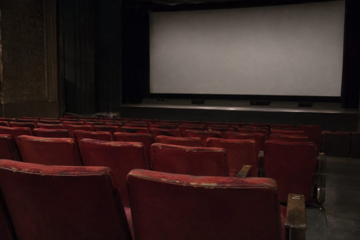 Rows of empty red seats in a darkened movie theater auditorium, screen blank and lit only by house lights.
