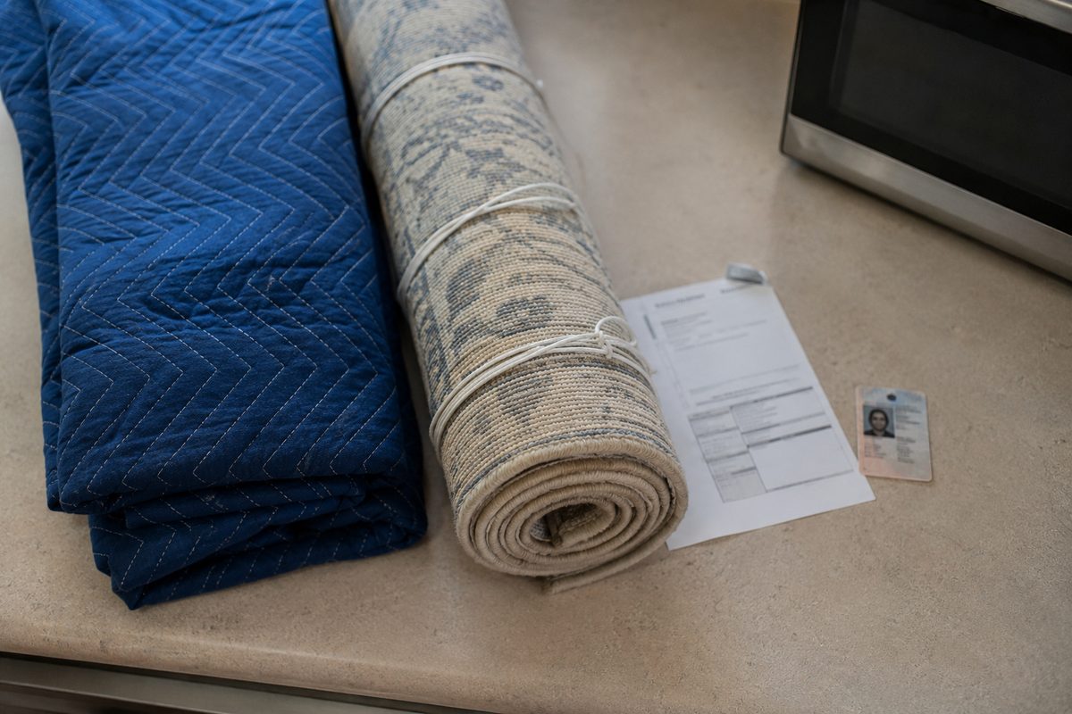 Photo illustration: stylized flat editorial vector, overhead flat-lay of a folded moving blanket, a rolled and tied carpet, a utility bill and a driver's license laid side by side on a plain counter, one small appliance pushed outside the frame's edge; neutral daylight, no people, no logos, no agency seals.