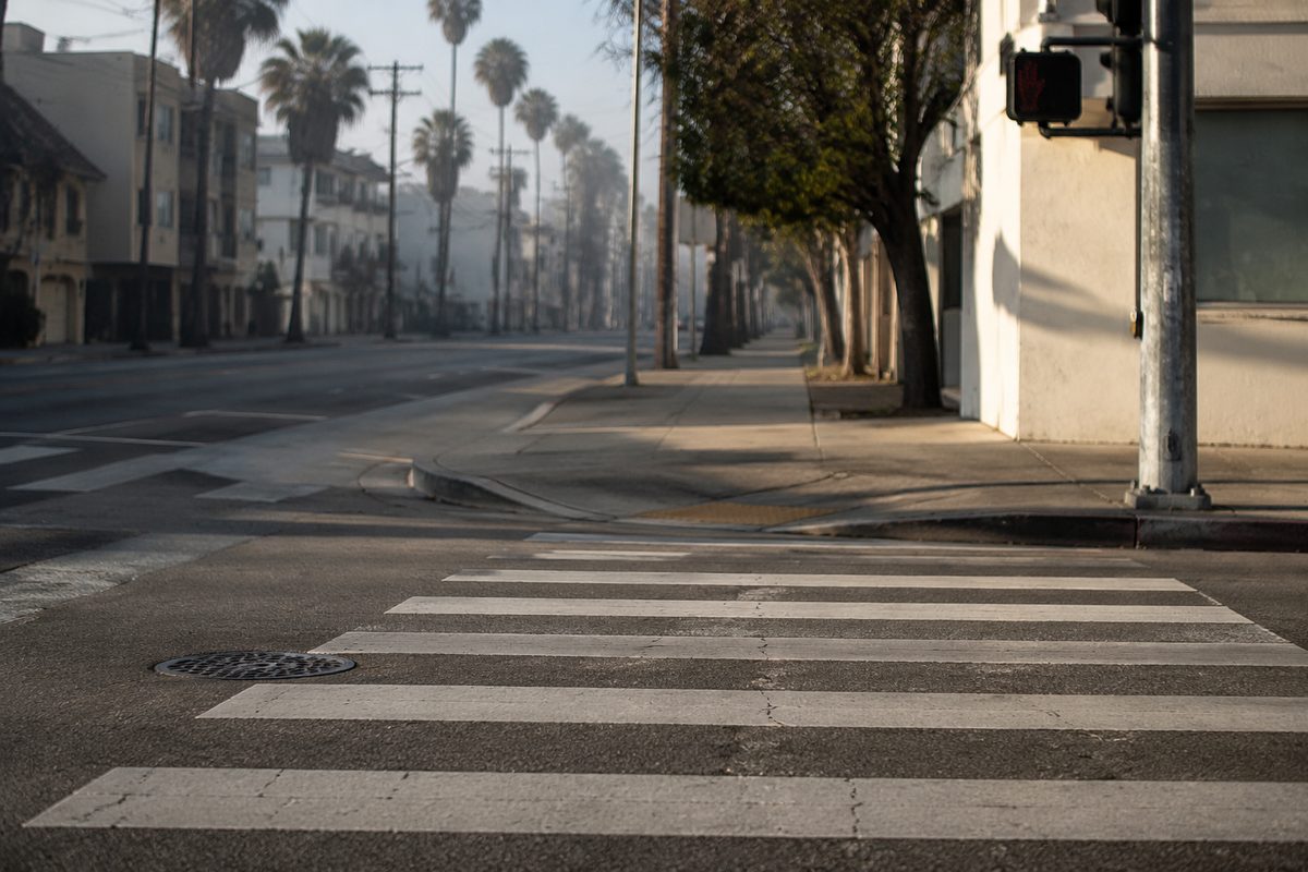 Photo illustration: a photo-illustration of an eye-level urban street corner in Los Angeles — a painted crosswalk in the foreground, no vehicles parked in the twenty feet closest to it, a faded curb (no red paint), no readable business signage, morning marine-layer light, no people identifiable. Handbook Desk service photography, per the desk's art card.