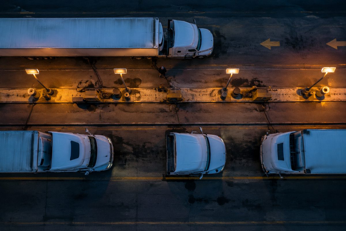 Overhead view of tractor-trailers lined up at a truck stop fueling island at dusk under orange lights.
