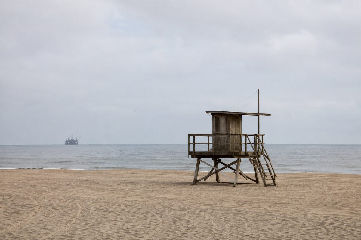 A wide, empty Southern California beach at midday with an unstaffed lifeguard tower and the faint silhouette of an offshore platform on the horizon
