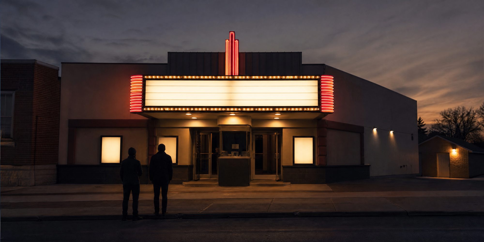 A small movie theater facade at dusk, shot straight on, its blank marquee lit and its letters unreadable, with two silhouetted figures at the curb.