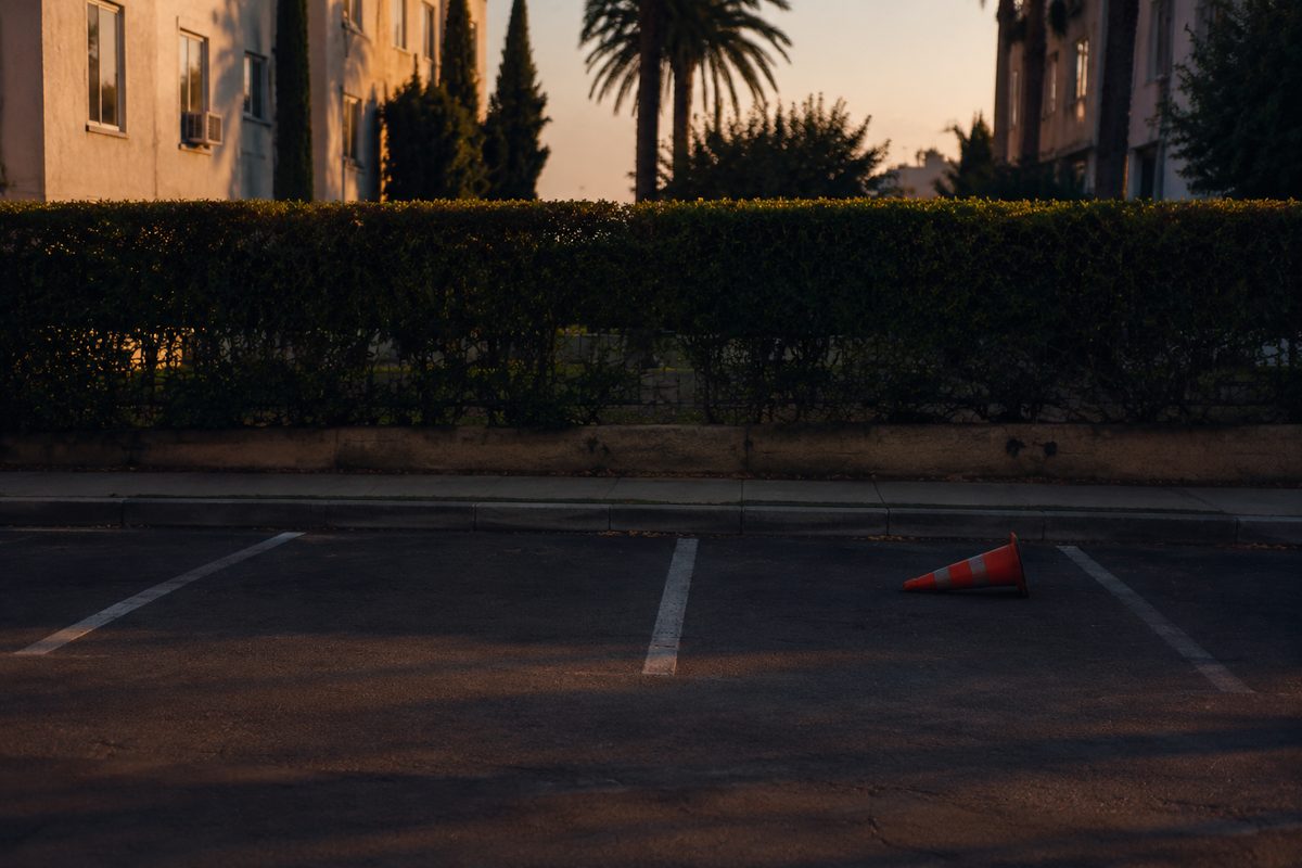 Photo illustration: an empty parking space on a residential Los Angeles street at dusk, painted stall lines and a curb, one orange traffic cone knocked sideways, no cars, no people, no signage or business names, long shadows