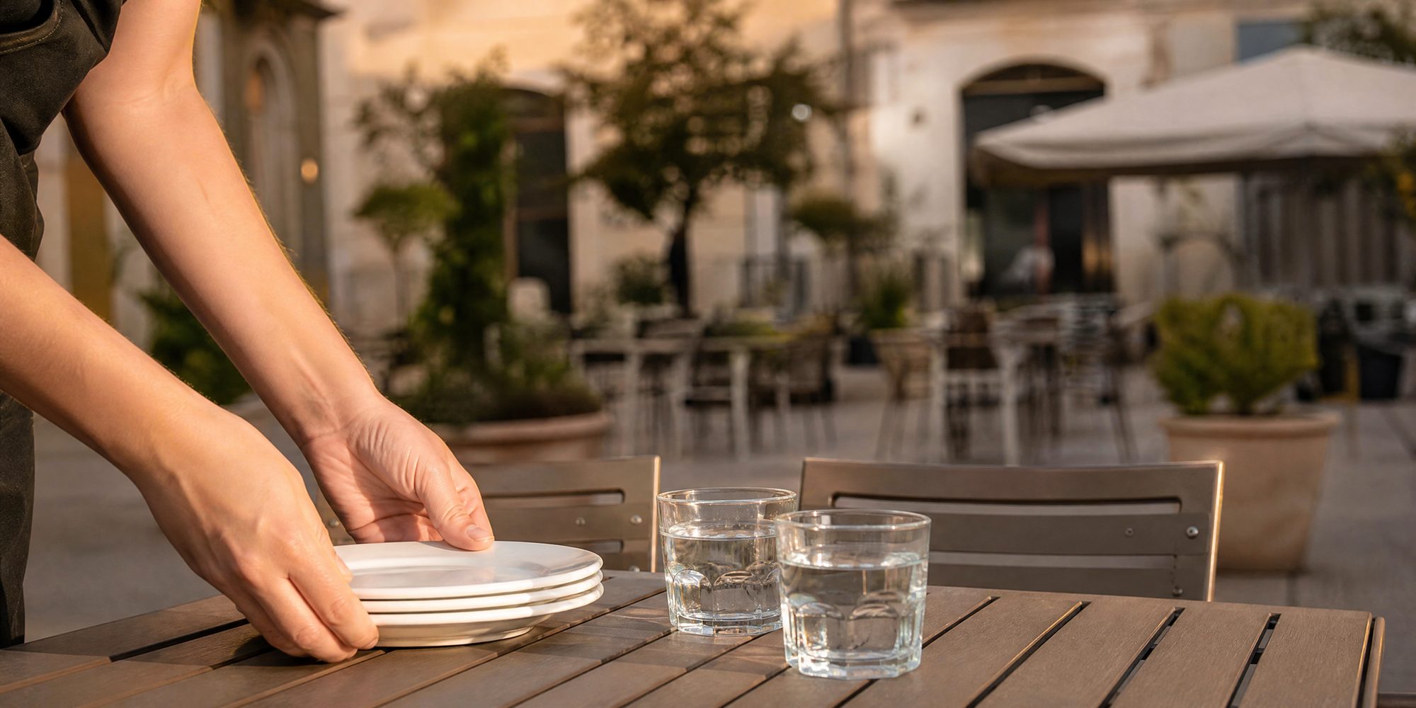 Two hands setting stacked white plates and water glasses on an outdoor cafe table at golden hour, an open-air courtyard blurred behind.