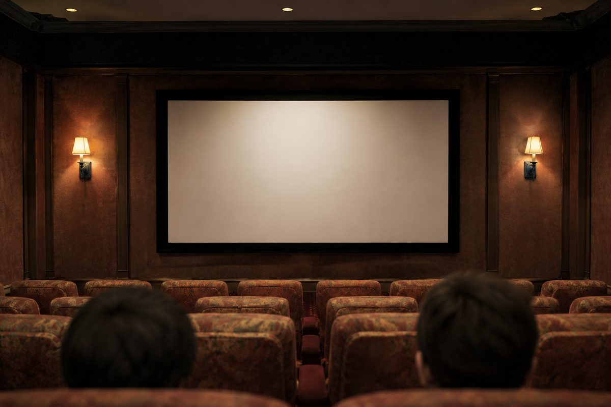 Symmetrical view of a small screening room: blank projection screen, tungsten wall sconces, and the backs of two heads in the front row.